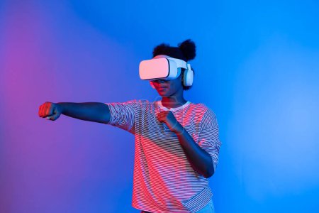 Smiling African American woman with VR headset playing boxing game turning to metaverse at vibrant blue and pink stadium portrait neon lighting in gaming technology interface 3D graphic. Contrivance.