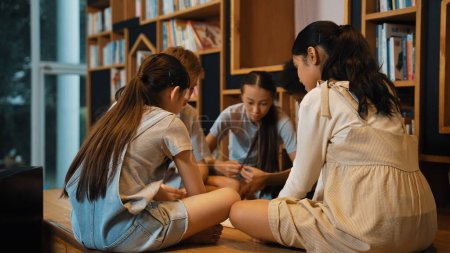 Photo for Group of diverse children sitting in circle while reading a book at library. Highschool girl sharing a magazine while pointing at interested topic and talking with multicultural friends. Edification. - Royalty Free Image