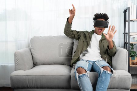 Photo for Surprised African American looking VR enjoy watching by touching the air in fantastic 3D cartoon movie technology virtual reality metaverse journey new world at living room sitting sofa. Contrivance. - Royalty Free Image