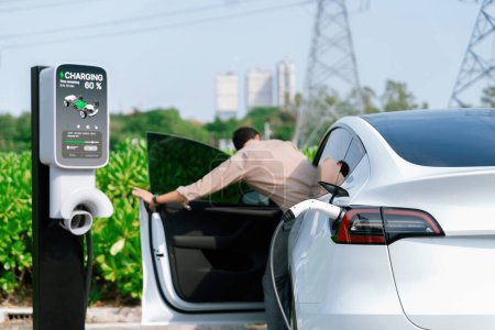 Young man recharge EV car battery at charging station connected to power grid tower electrical industrial facility as electrical industry for eco friendly vehicle utilization. Expedient