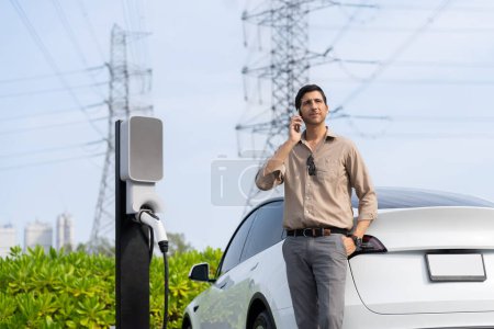 Téléchargez les photos : Homme parlant sur le téléphone tout en rechargeant la batterie de voiture EV à la station de charge connectée à la tour de réseau électrique comme industrie électrique pour une utilisation de voiture respectueuse de l'environnement. - en image libre de droit