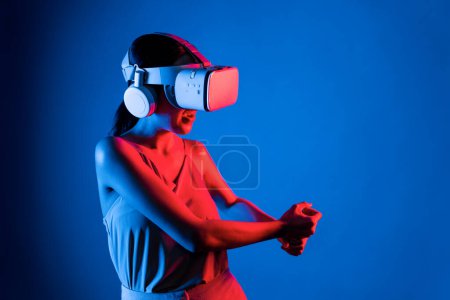Téléchargez les photos : Smart femelle stand porter casque VR connexion metaverse, future technologie de la communauté cyberespace. Femme élégante à l'aide de mains contrôlant sabre laser virtuel sérieusement jouer à des jeux de combat. Hallucination. - en image libre de droit