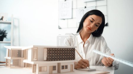 Closeup image of young architect engineer writing house structure in blueprint with document and architectural model placed on table at office. Creative design and architect concept. Immaculate.