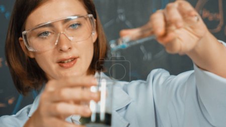 Photo for Closeup of young beautiful teacher focus on mixing chemical liquid at laboratory. Skilled scientist doing an experiment by inspect colored solution while standing in front of blackboard. Erudition. - Royalty Free Image