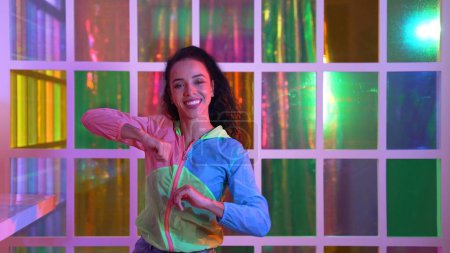 Photo for Happy hispanic girl in lively mood moving along music in neon light while wearing colorful cloth at night club. Street dancer making energetic footstep or movement while looking at camera. Regalement. - Royalty Free Image