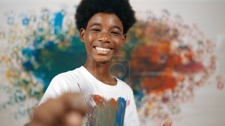Photo for African boy standing at colorful stained wall while holding brush and point at camera. Happy highschool student wearing white shirt with stain color and look at camera with confident. Edification. - Royalty Free Image