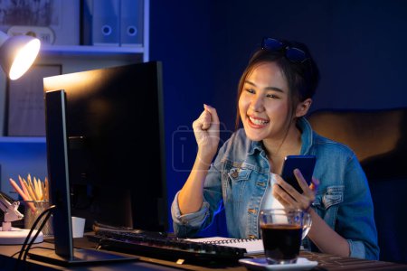 Sonriendo joven hermosa oficial creativa asiática mirando en PC en el objetivo de ventas en línea, mientras que la celebración de la taza de café en la sala de luz de neón en la noche, ganando puestos de trabajo freelancer estrategia de marketing. Estratagema.