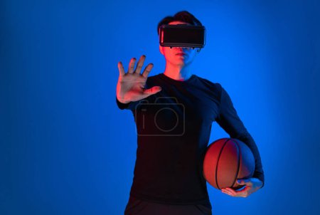 Téléchargez les photos : Athlète asiatique avec VR tenant un joueur de basket-ball explorant l'espace virtuel sur la formation sportive portrait néon reliant numérique technologie hologramme futuriste métaverse terrain de jeu du monde. Contrivance. - en image libre de droit