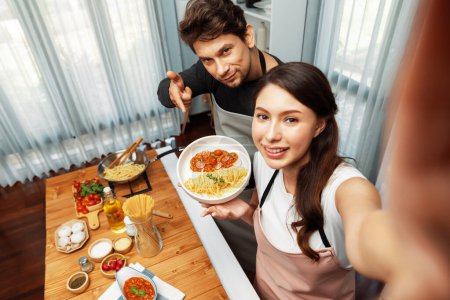 Hacer foto selfie en pareja influencers chef espaguetis completamente cocidos con carne rematada con salsa de tomate grabación de plato especial en cámara, sirviendo comida saludable en el chanel casero moderno. Postulado.
