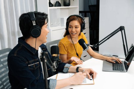Smiling radio influencer hosts wearing headphones with channel social media online live streaming with talking topic on script note and laptop to listeners at morning time at studio record. Infobahn.