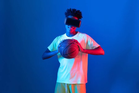 Téléchargez les photos : Jeune joueur afro-américain de basket-ball portant VR rebondissant pose sur la lumière rose bleu néon connectant la technologie numérique futuriste réalité virtuelle à Metaverse World Play Court. Contrivance. - en image libre de droit