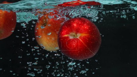 Eine Nahaufnahme einer frischen lebendigen roten Tomate, die in klarem Wasser mit schwarzem Hintergrund schwimmt. Makrographie eines Bio-Apfels, der fällt und ins plätschernde kühle Wasser fällt. Frische-Konzept. Pabulum.