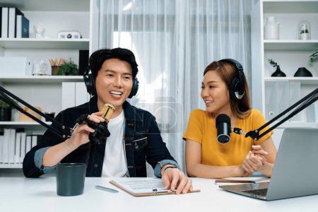 Smiling radio influencer hosts wearing headphones with channel social media online live streaming with talking topic on script note and laptop to listeners at morning time at studio record. Infobahn.