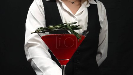Macrography, witness the artistry of a bartender serving a vibrant red cocktail in a martini glass, beautifully adorned with a sprig of fresh rosemary against a sleek black background. Comestible.