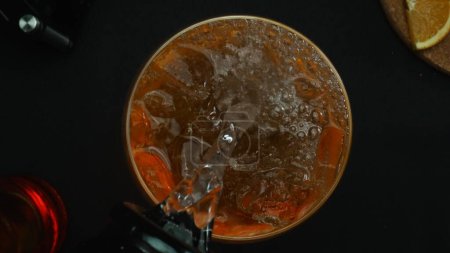Macrography, experience the art of mixing a Negroni cocktail from a top-down perspective, adorned with a fresh vibrant orange slice, all against a dramatic black background. Top down view. Comestible.