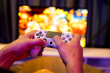 Close up one hands man player of videospiel on TV mit Joystick-Steuerung mit verschwommenen Kampf-Gaming-Bildschirm mit Snack und Getränken bei dekorativer mit Neon-Blaulicht im gemütlichen Wohnzimmer. Postulat.