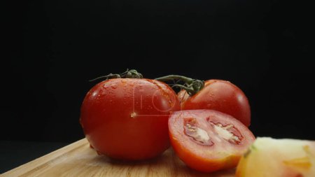 Macrographie, tranches de tomate reposent élégamment sur une planche à découper rustique sur un fond noir dramatique. Chaque gros plan capture la texture juteuse et les couleurs riches des tomates. Comestible.