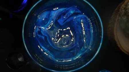 Macrography with a top-down view of a professional bartender hand elegantly serving a Blue Hawaii cocktail with black background. Close-up shot captures the fluid motion. Aerial view. Comestible.